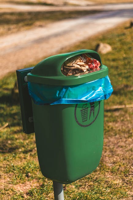 A green outdoor waste bin with a black lid and a light blue flap on top is situated on a grassy area within a garden setting. The bin features a white icon of a person disposing of trash into a bin, e